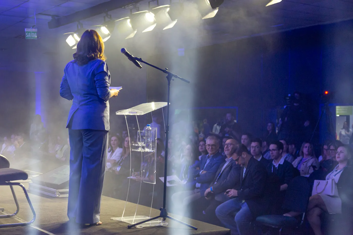Sonia Campos no palco, de costas, diante do público de um congresso